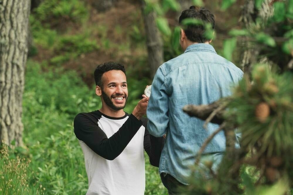 Mikah is down on one knee, holding a ring box up to Sebastian, who is standing and facing Mikah. Mikah is smiling brightly, wearing a black and white long-sleeve shirt, while Sebastian, in a light blue denim shirt, looks down at Mikah. They are in a green forested area.