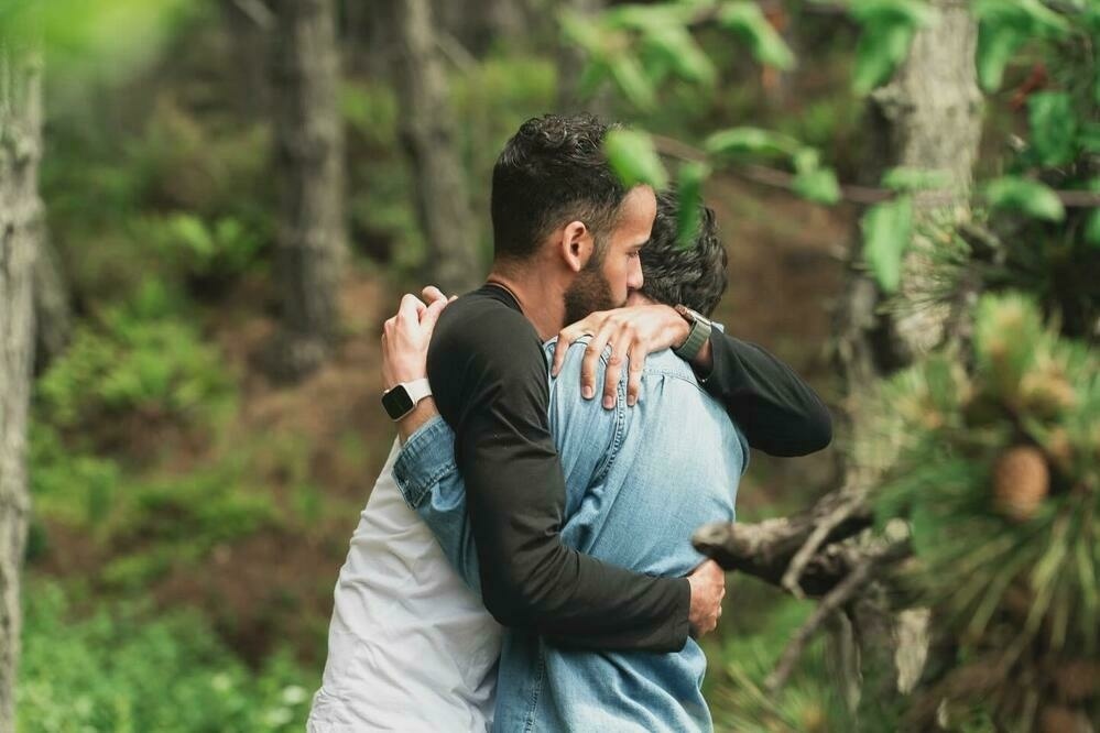 Mikah and Sebastian share a warm embrace in a wooded area. Their faces are close together, showing a tender moment.