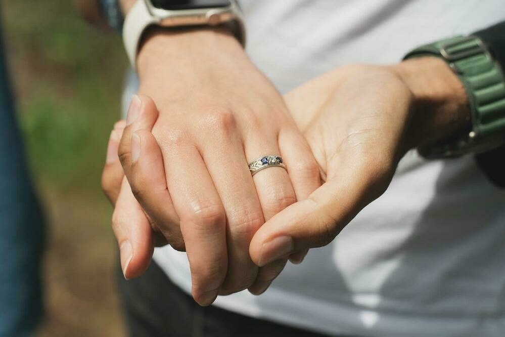 A close-up of two hands holding each other. One hand, belonging to Mikah, wears a green wristwatch, while the other hand, belonging to Sebastian, wears a white gold ring with two sapphires and a diamond.