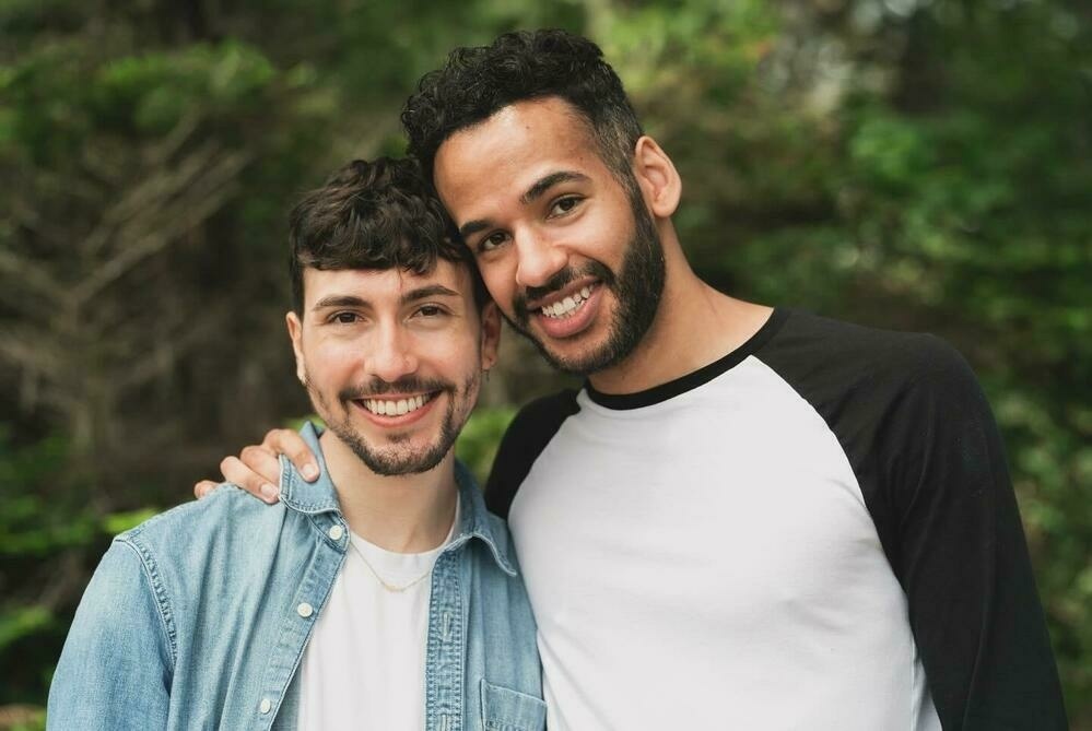 Mikah, wearing a black and white long-sleeve shirt, stands next to Sebastian who is wearing a light blue denim shirt. They are both smiling warmly and standing close together, with Mikah's arm around Sebastian's shoulder. The background is a green, leafy outdoor setting.