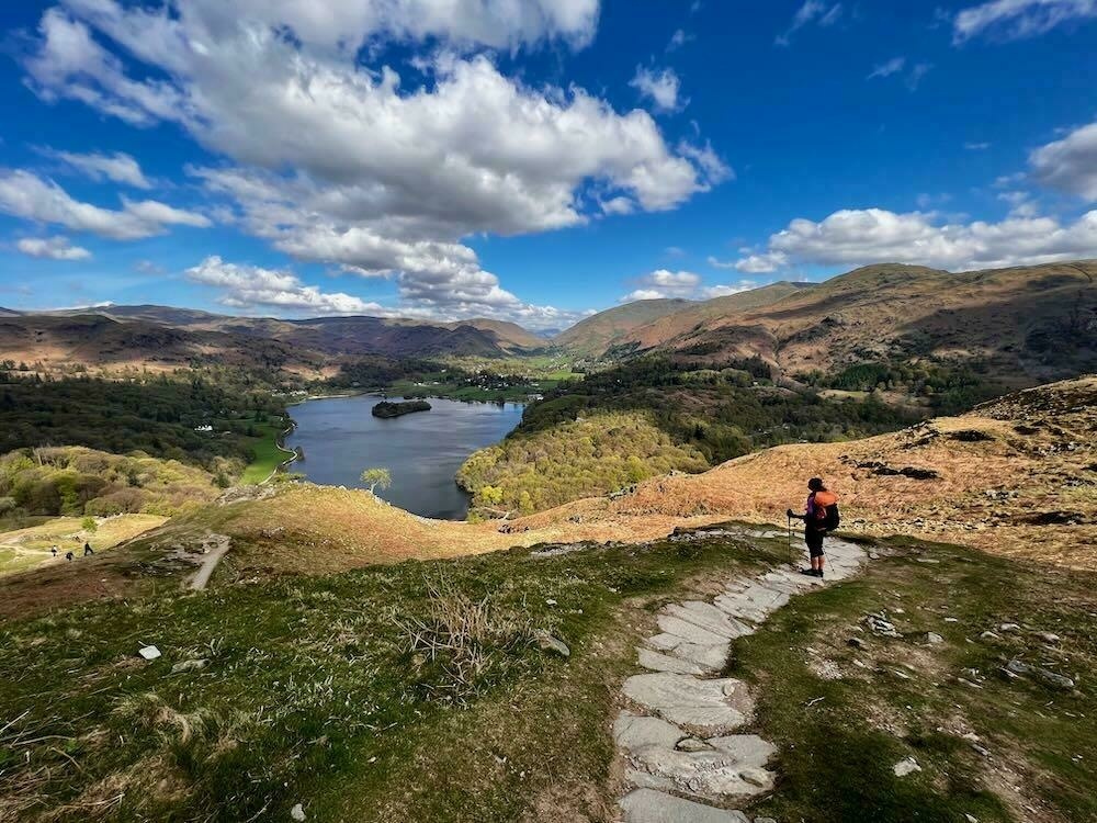 A woman stands above Grasmere water with a giant backpack on.