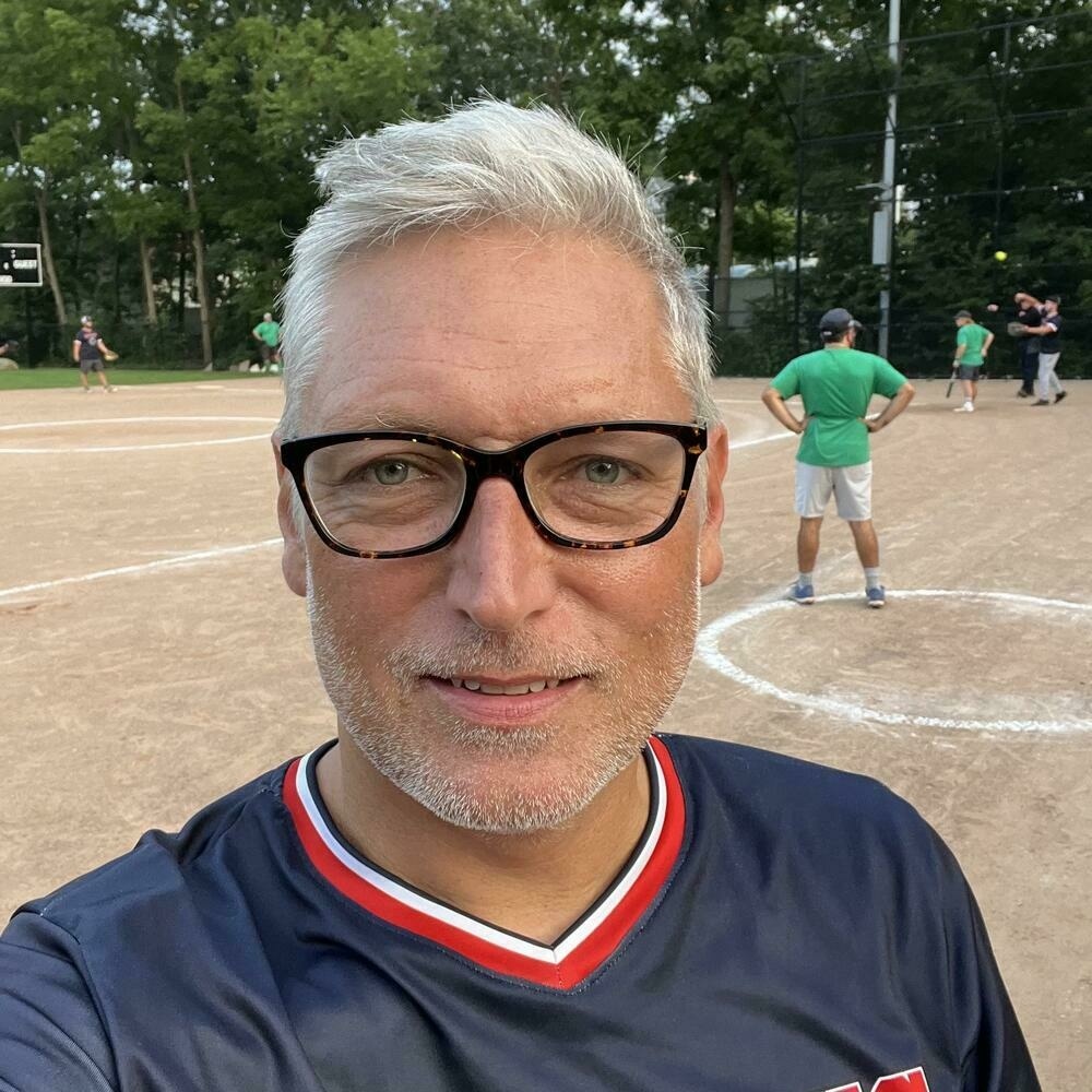 Selfie of a man with tan caucasian skin, a white stubble beard, and white/grey hair. Wearing glasses, wearing a blue baseball jersey and looking right at the camera. Baseball diamond in the background.