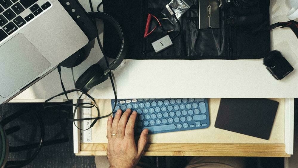 A person is working on a blue keyboard at a cluttered desk with various tech gadgets and accessories.