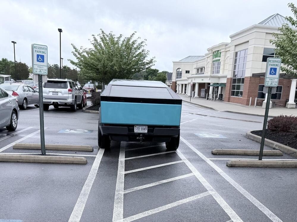 A cybertruck with a wrap parked in the area between handicapped parking spots, where nobody is supposed to park. 