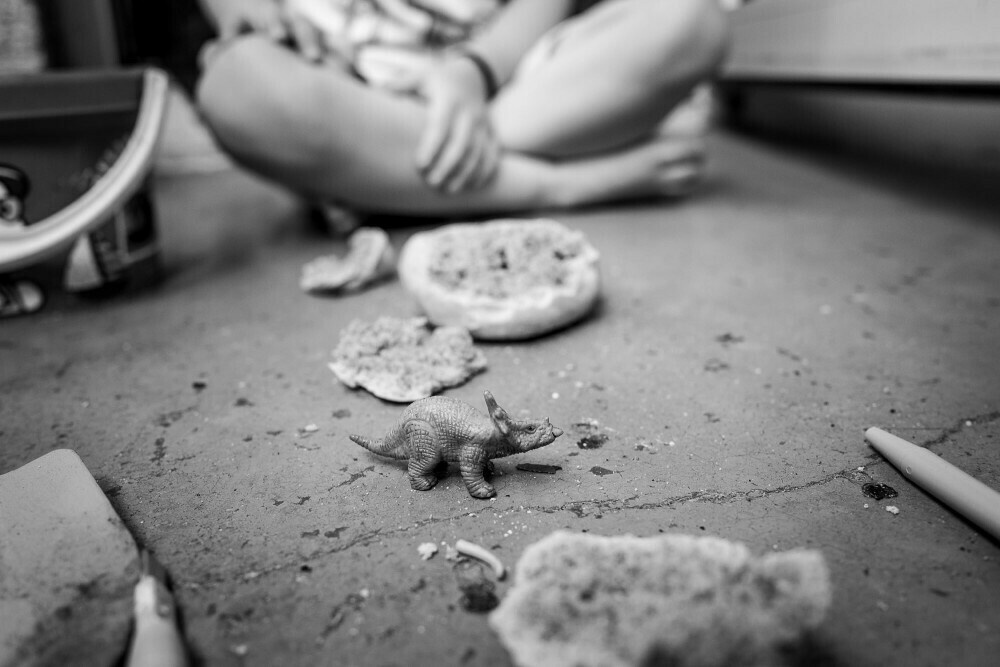 A little dinosaur figurine sits on the ground next to remants of dinosaure egg shells made of salt dough