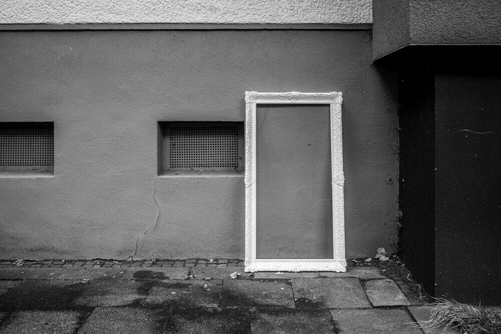 Monochrome image of a white frame leaning against a wall on a grimy sidewalk