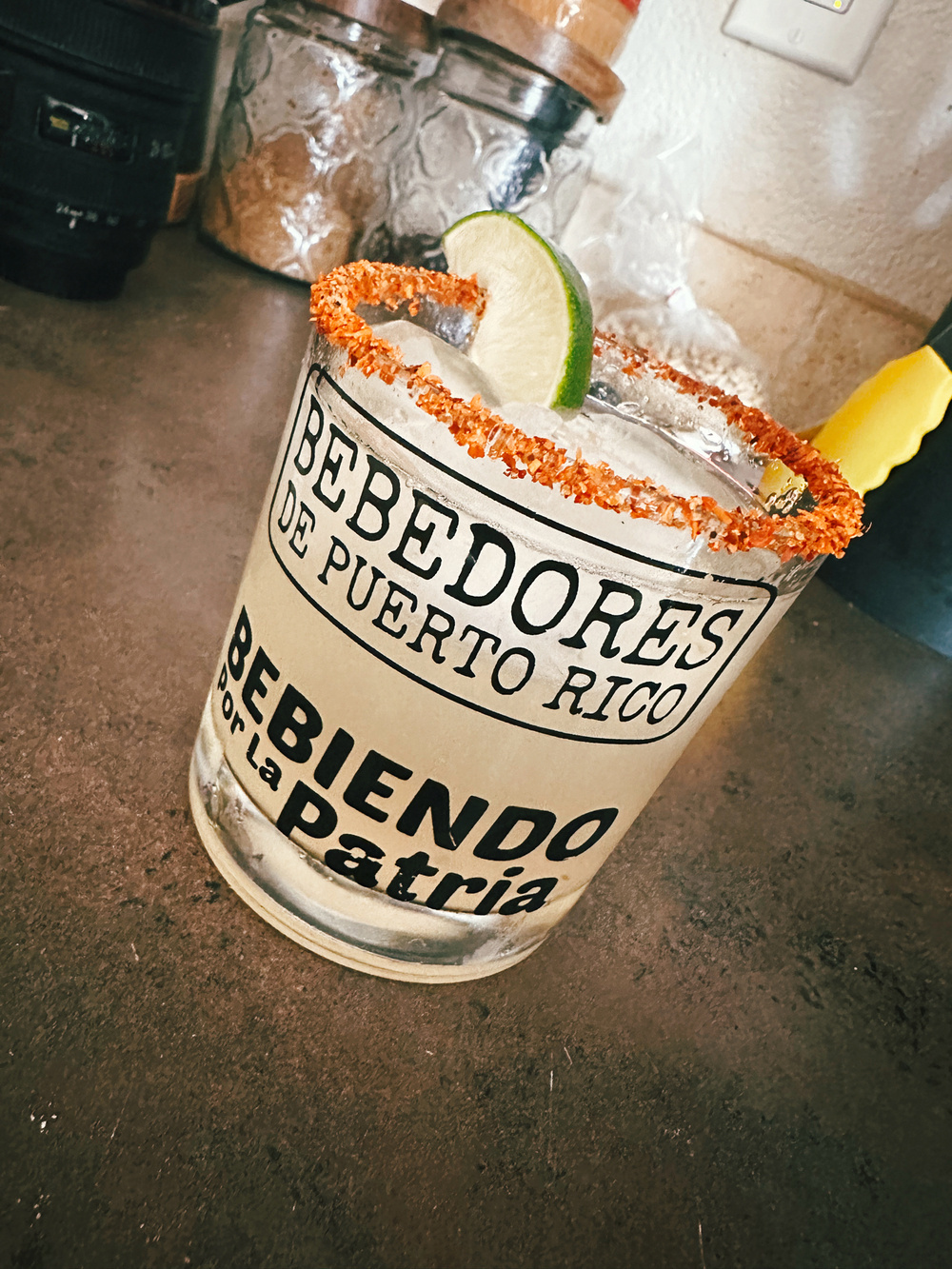 A cocktail in a glass with a chili-lime rim and a lime wedge garnish. The glass has text reading “BEBEDORES DE PUERTO RICO” and “BEBIENDO por La Patria.” 