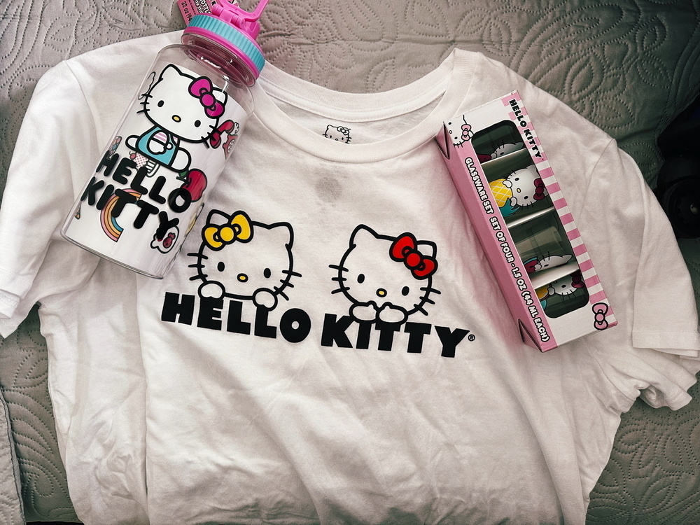 This image shows a collection of Hello Kitty merchandise, including a white T-shirt with three Hello Kitty faces and the text “HELLO KITTY,” a Hello Kitty water bottle with a pink lid, and a boxed set of four Hello Kitty shot glasses 