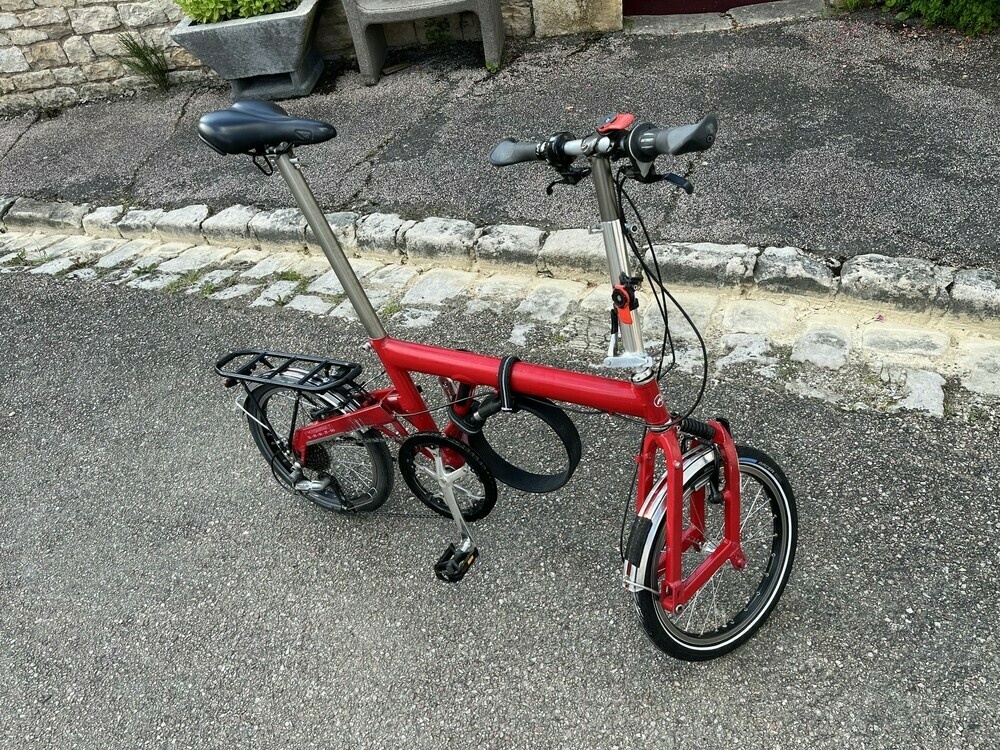 Red Birdy bike with back rims and silver mudguards. It stands on tarmac with a stone curb behind. 