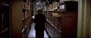 gif of librarian from Ghostbusters walking down an aisle with books on the left and a large card catalog on the right. As she walks down drawers begin opening on their own and cards begin flying up into the air and swirling around.