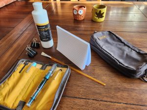 Wooden table featuring a deck index cards bound by glue at the top stand up near some PVA adhesive surrounded by some binder clips, a paint brush, a Lochby case of fountain pens and a stationery bag.