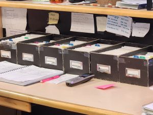 Six well-used, beat up, and taped together brown cardboard card index boxes filled with index cards and sporadically interspersed with alphabetic card dividers sitting on a desk with a variety of notes and Post-Its stuck to the wall behind it