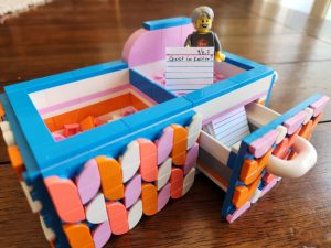 What appears to be a small two drawer zettelkasten made out of LEGO in a somewhat garrish blue, white, orange and pink color scheme. The right drawer is open with a few dozen mini-index cards inside. On top is a LEGO figurine with gray hair, a beard. and mustache. It is holding a tiny index card that reads "9/8,3 Geist im Kasten?"