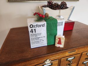 A brick of 10 packs of 100 index cards boldly labeled Oxford 41. They are wrapped with a green ribbon and a sprig of evergreen with red berries and a small Santa Claus card all sitting on a wooden card index.