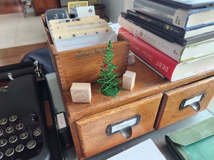 My desk today featuring a few card indexes, a pile of books and a typewriter. In the center of the photo is a small, green metal ornament in the shape of a small Christmas tree with tiny red decorative balls.