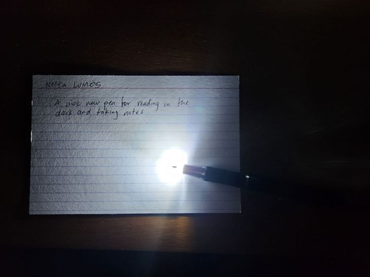Taken in a dark room, an index card is partially illuminated from the tip of a ballpoint pen which has an embedded LED light. Handwritten on the card is the title "Nota Lumos" with the phrase "A nice new pen for reading in the dark and taking notes."
