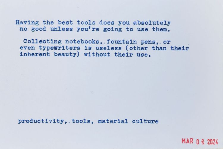 Typewritten blue text on a blank index card reads: Having the best tools does you absolutely no good unless you're going to use them. Collecting notebooks, fountain pens, or even typewriters is useless (other than their inherent beauty) without their use. productivity, tools, material culture