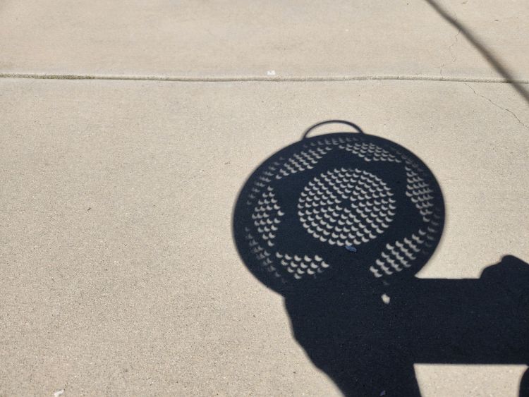 Shadow of a colander held up in the sunshine at the peak of the eclipse. The multi-pinhole camera shows a flower pattern of sun circles with a small bite taken out of each of them by the occlusion of the light by the moon all focused onto a concrete background.