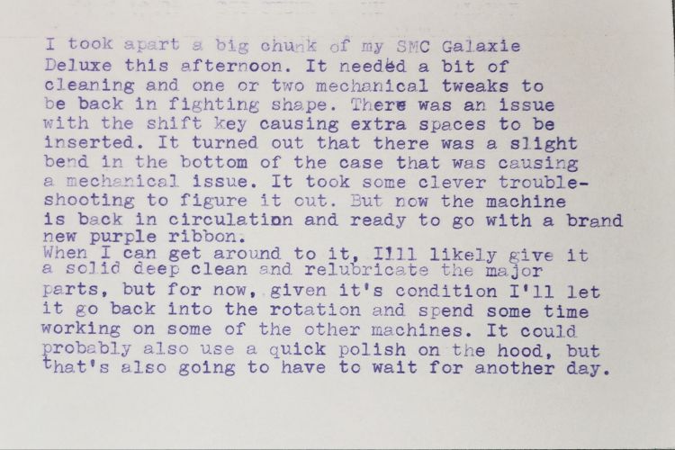 A typed white index card with purple ink which reads: I took apart a big chunk of my SMC Galaxie Deluxe this afternoon. It needed a bit of cleaning and one or two mechanical tweaks to be back in fighting shape. There was an issue with the shift key causing extra spaces to be inserted. It turned out that there was a slight bend in the bottom of the case that was causing a mechanical issue. It took some clever trouble- shooting to figure it out. But now the machine is back in circulation and ready to go with a brand new purple ribbon. When I can get around to it, I'll likely give it a solid deep clean and relubricate the major parts, but for now, given it's condition I'll let it go back into the rotation and spend some time working on some of the other machines. It could probably also use a quick polish on the hood, but that's also going to have to wait for another day.