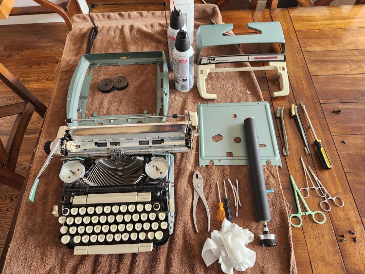 Photo down on a wooden table covered with a brown towel on which sits a somewhat dissassembled green Smith-Corona typewriter. The case has been removed into three parts as well as the platen. Strewn around it are a variety of pliers, screwdrivers, the platen, hemostats, and screws.