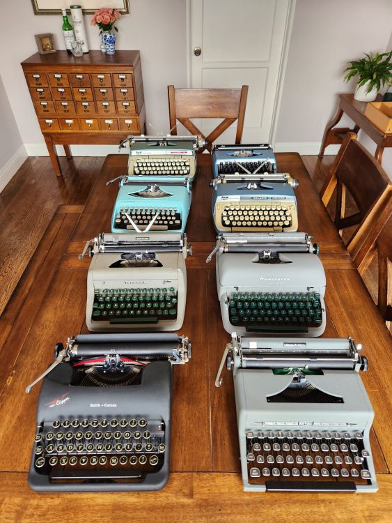 Two rows of typewriters lined up on a wooden table. From front to back, left to right are a curvy black '48 Smith-Corona Clipper with glass keys, a '49 Grey boxy Royal Quiet De Luxe with tombstone glass keys, a dirty '55 Royal Quiet De Luxe with green keys, a gray '57 Remington Rand Quiet-Riter with green keys, a bright 1960s teal Remington Streamliner, a blue and cream colored late 60s Smith-Corona Galaxie Deluxe, a green early 70s Smith-Corona Classic 12, and a low slung 70s blue metal Brother Correction 11.