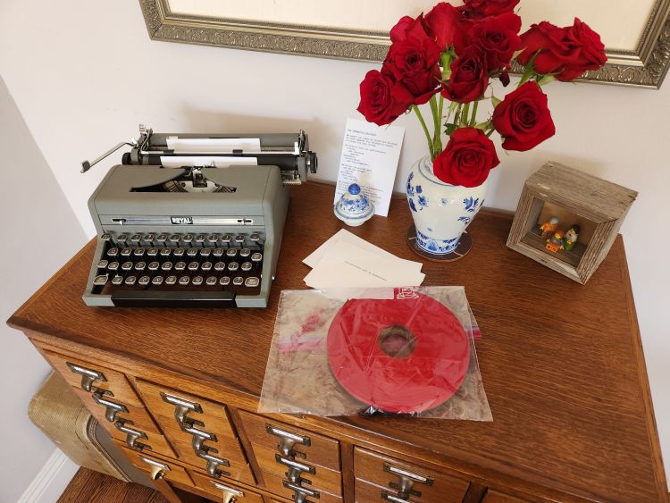 A full reel of red and black typewriter ribbon in a clear plastic bag with some internal ink smudges sits on a 20 drawer library card catalog next to a Henry Dreyfuss Royal Quiet De Luxe typewriter and a vase full of red roses.