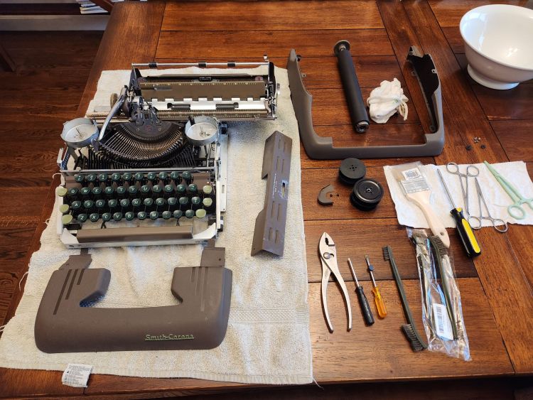 A deconstructed typewriter sitting on a towel on a wooden table surrounded by the brown steel body portions, screws, typewriter ribbon reels, screwdrivers, pliers, and brushes.