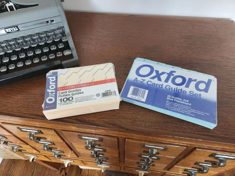 Two packages of index card guides sitting on a library card catalog next to a gray Royal typewriter. The manilla package has 4x6" guides and the 5x8" guides are in blue pressboard.