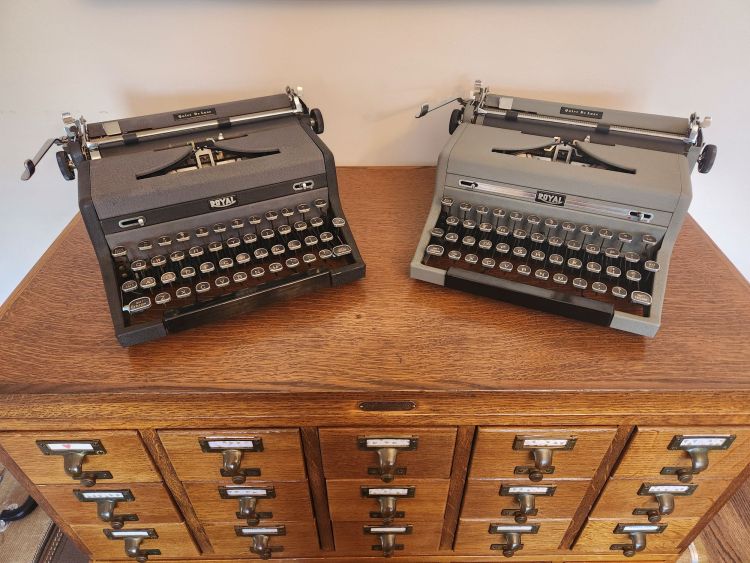 Two Royal Quiet De Luxe typewriters sitting on top of a library card catalog at angles to each other. One on the left is black with grey trim while the other is gray with chrome trim.