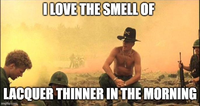 Screencapture from the film Apocalypse Now featuring a shirtless man with a calvary-style military hat squatting down on a beach while two soldiers work in front of him. He is literally surrounded by the yellow fog of war. The meme image is surrounded with the words: I love the smell of lacquer thinner in the morning.