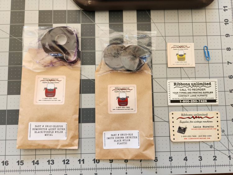Contents of an order from Ribbons Unlimited laid out on a crafting mat. On the left are a pair of Remington metal hubs with black and purple ribbon. To the right are a pair of small spools for a Skyriter with black ribbon and next to this are business cards and stickers from the company.