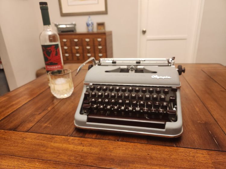 A light gray Olympia SM3 De Luxe typewriter on a wooden table next to a highball glass of Penderyn whisky. In the background we can see a library card catalog.