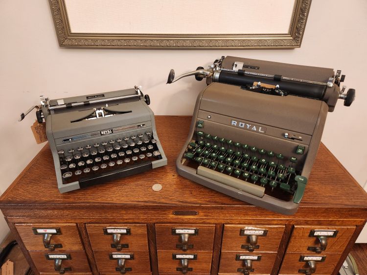 A straight on view of a small gray Royal QDL typewriter next to a brown Royal HH which is almost 2 1/2 times the size of the first. Both sit on top of a wooden library card catalog.