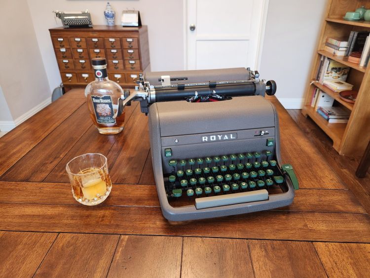 A crystal old fashioned glass of bourbon on the rocks next to a Royal HH standard typewriter