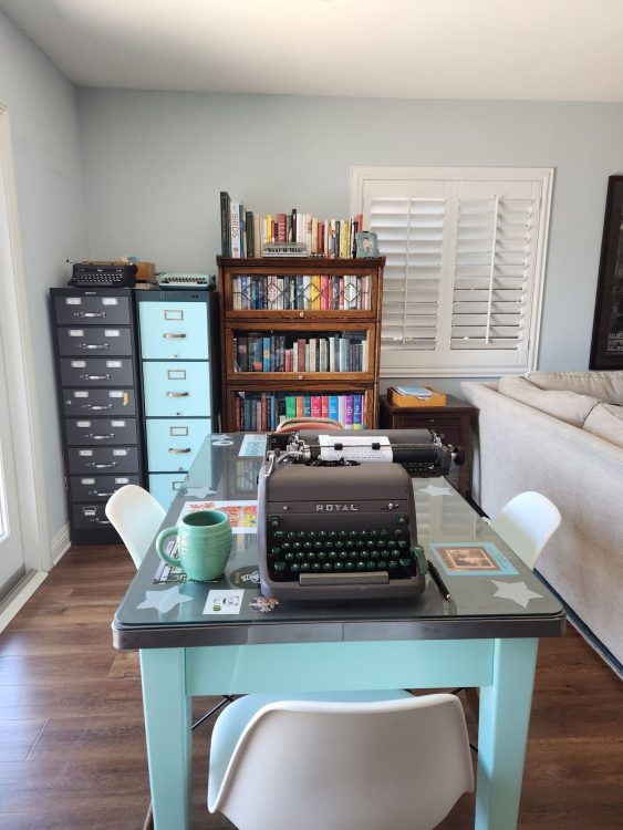 Office setting with a green metal table in the foreground with a brown Royal HH typewriter on it. In the background are a card catalog filing cabinet and a four drawer filing cabinet whose paint matches the table. There's also a barrister bookcase full of books.