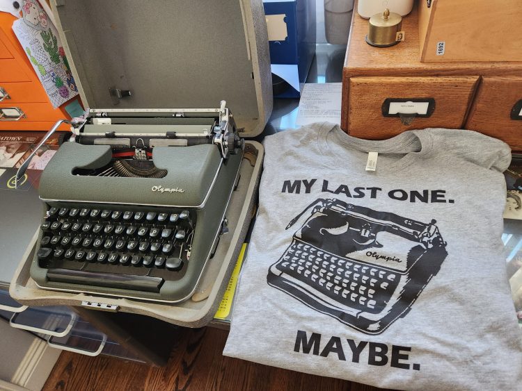 Sitting on a desk are a green Olympia SM3 typewriter in its case next to a gray t-shirt with a black and white image of the same typewriter surrounded with the text: "My Last One. Maybe"