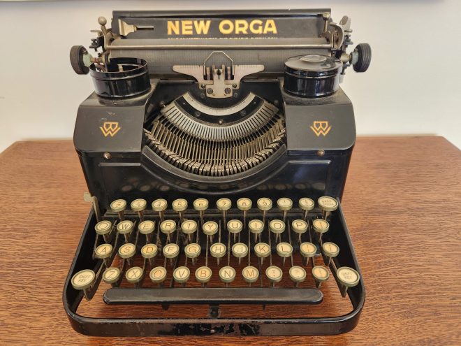 Frontal view of a black 1930s typewriter with a bold gold name New Orga on the paper table. The black enamel typewriter has glass keys with yellowed key legends.