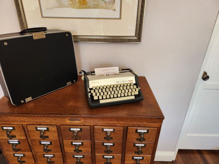 A black and cream Remington 666 typewriter next to ita black case sits on a wooden library card catalog