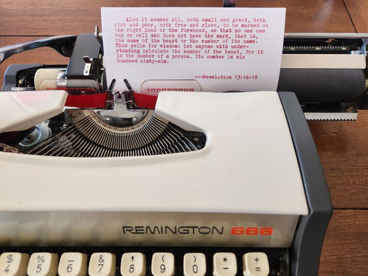 The carriage of a Remington 666 Typewriter down to the first row of number keys. Sitting in the platen is a white index card with red typewritten text above a bright red monochrome ribbon. It contains a quote from Revelation 13:16-18 which reads: Also it causes all, both small and great, both rich and poor, both free and slave to be marked on the right hand or the forehead, so that no one can buy or sell who does not have the mark, that is, the name of the beast or the number of its name. This calls for wisdom: let anyone with understanding calculate the number of the beast, for it is the number of a person. Its number is six hundred sixty-six.