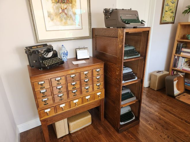 A Fred Macey Company 20-drawer card index filing cabinet with the drawers and hardware removed. In their place we see four typewriters peeking out from each of four shelves inside and an additional typewriter on top.  Next to the filing cabinet is a Gaylord library card catalog with a black typewriter on top along with some additional decorations.