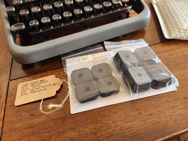 Four black rubber feet in a clear plastic bag next to four black rubber spacers in another similar bag sitting in font of the keyboard of a typewriter with a gray body. Off to the side is a brown identification tag with typed details about the typewriter.