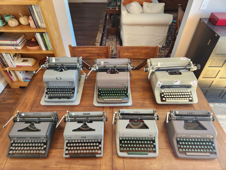 Two rows of primarily gray and brown vintage typewriters on a wooden dining table.