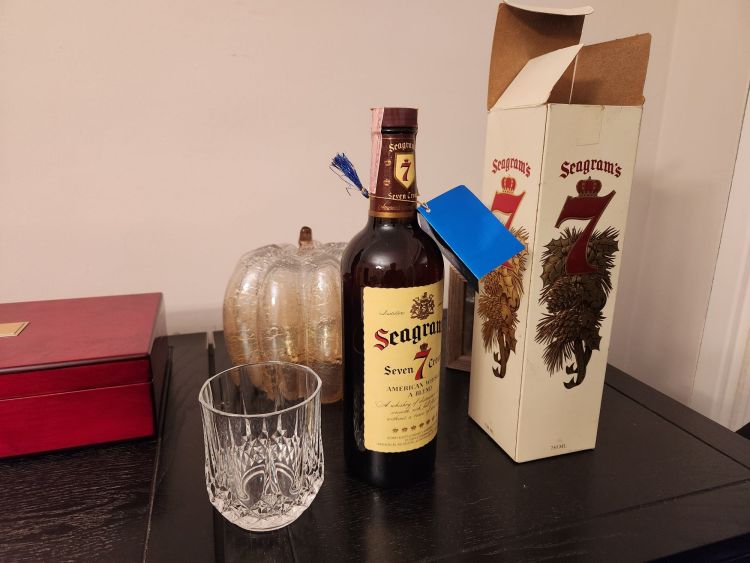 A black bar top with a crystal old fashioned glass next to an unopened bottle of Seagrams 7 and its gift box.