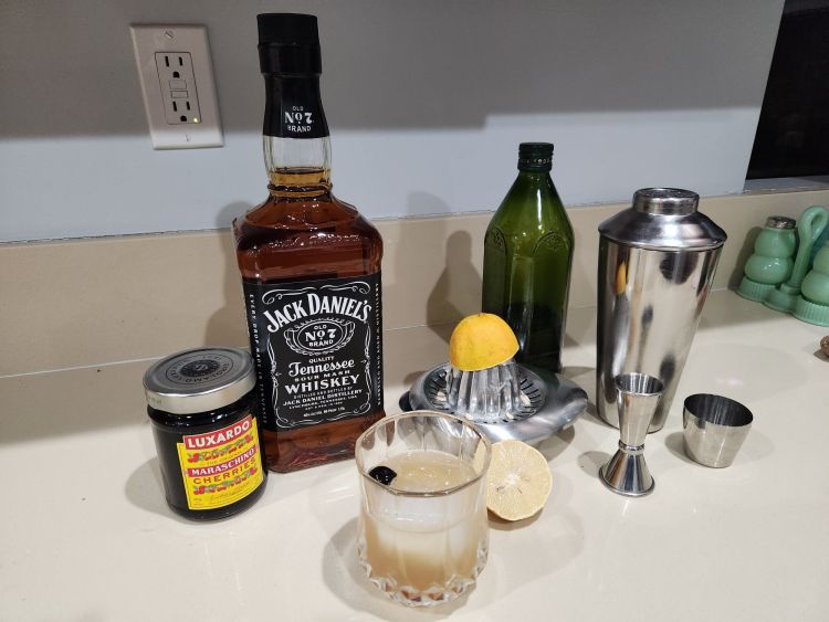 Counter with the accoutrement for making a whiskey sour including a bottle of Jack Daniels, a jar of Luxardo cherries, a metal juicer with a lemon, a bottle of sugar syrup and a stainless steel shaker
