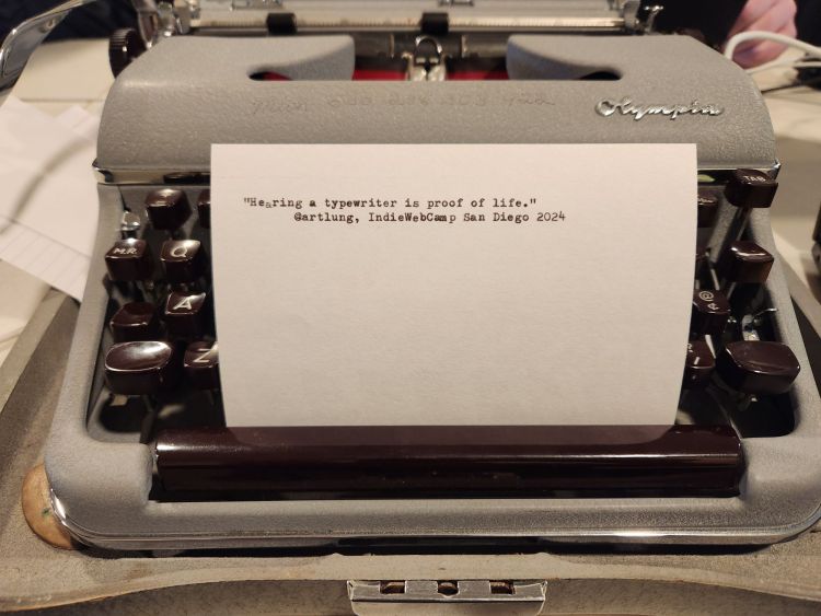 Typed index card propped up in the brown keys of a gray Olympia SM3 typewriter that reads: "Hearing a typewriter is proof of life." @artlung, IndieWebCamp San Diego 2024