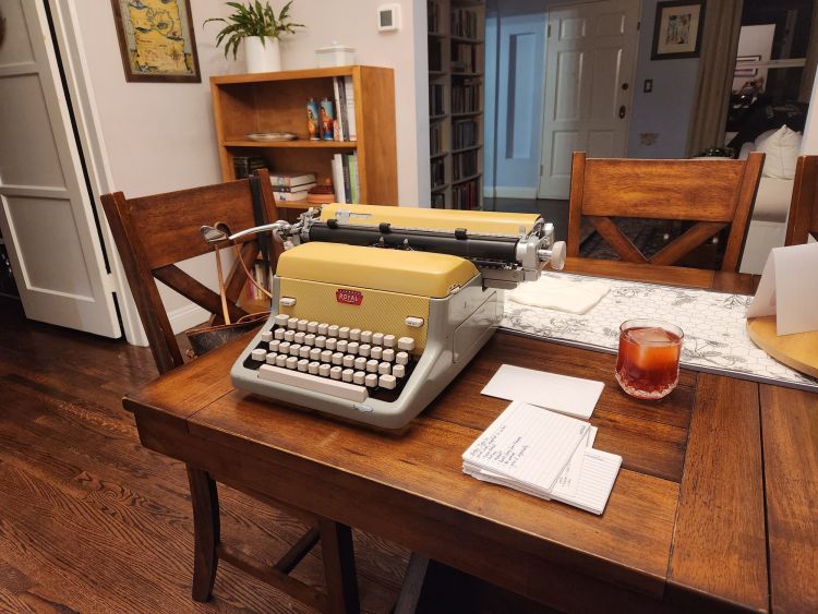 Yellow Royal FP typewriter on a wooden table next to some index card notes and a crystal glass containing a Negroni.