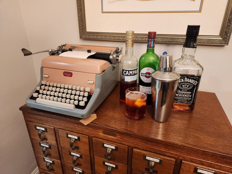 A double old fashioned with a redish looking Boulevardier cocktail in it surrounded by the bottles of the constituents and a Royal FP typewriter in pink all sitting on a library card catalog