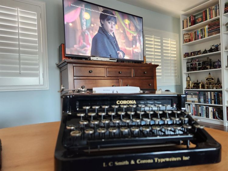 A black Corona flat top typewriter in the foreground with an index card reading "Wednesday Core" in it. In the background is a television playing season 2 of Wednesday on Netflix featuring Jenna Ortega as Wednesday Addams. It almost looks as if she's staring over to the Corona typewriter jealously.