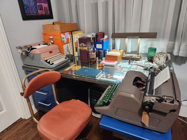 Office set up showing a gray executive tanker desk with blue drawers and writing drawers. In the front is a brown Royal HH standard typewriter with a visually similar vintage brown metal desk lamp with art deco styling behind it. Off to the side is a box for and bottle of Isle of Sky 18 year old whisky with a double old fashioned glass nearby.