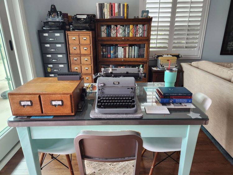 A bright room with a green steel table on top of which sits a 1950 gray Royal KMG typewriter, a two drawer Shaw-Walker oak card index and some papers and mathematics textbooks. Behind the table is a barrister's bookcase full of books, and two card index filing cabinets.